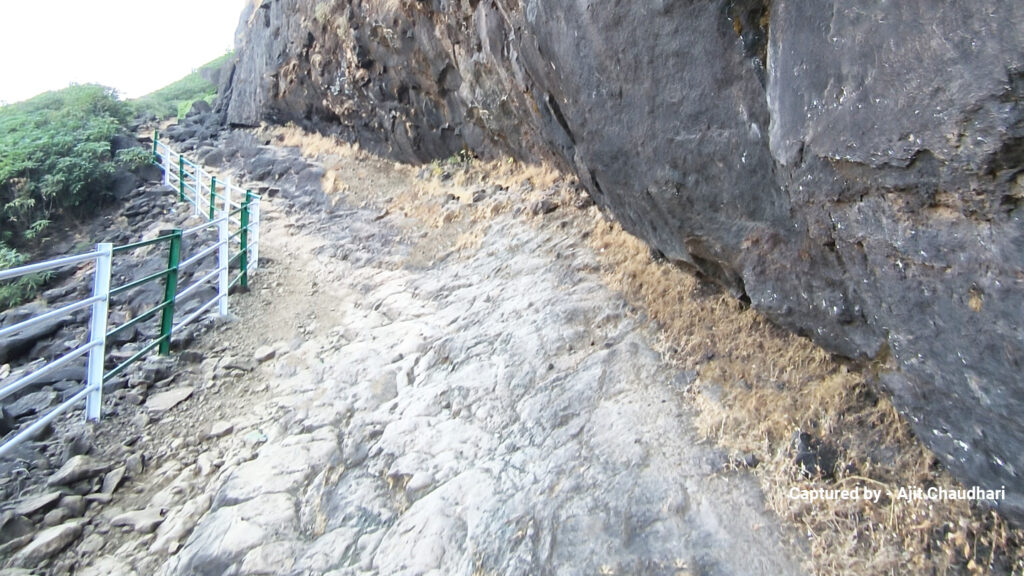 A rocky path curves around a steep cliffside, bordered by a metal railing for safety. The area is rugged and natural, conveying a sense of adventure.