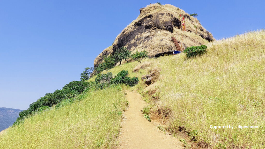 A narrow dirt path winds up a grassy hillside toward a rugged, rocky hilltop under a clear blue sky. Sparse green shrubs dot the landscape.