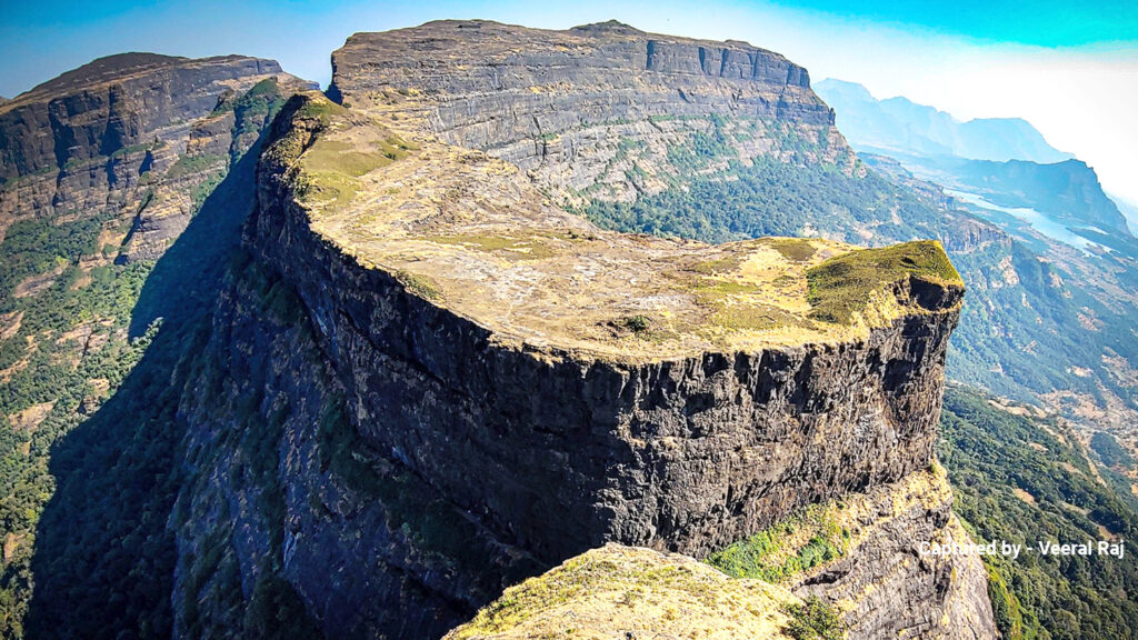 A majestic cliff with a flat, grassy top extends into the distance. Rocky edges contrast with lush greenery below, under a clear blue sky. Rugged and serene.