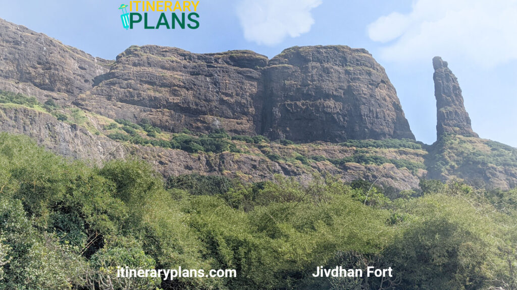 A scenic view of Jivdhan Fort, featuring rugged, towering rock formations against a clear blue sky. Dense green foliage covers the hillside below.