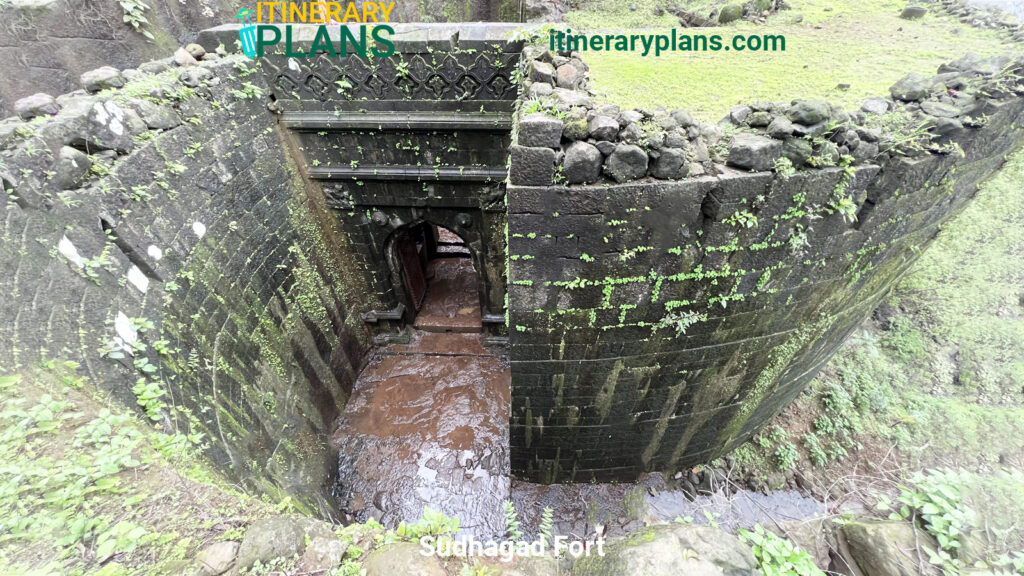 Ancient stone gateway of Sudhagad Fort, with moss-covered walls and a muddy path leading through. The scene conveys age and history amidst lush greenery.