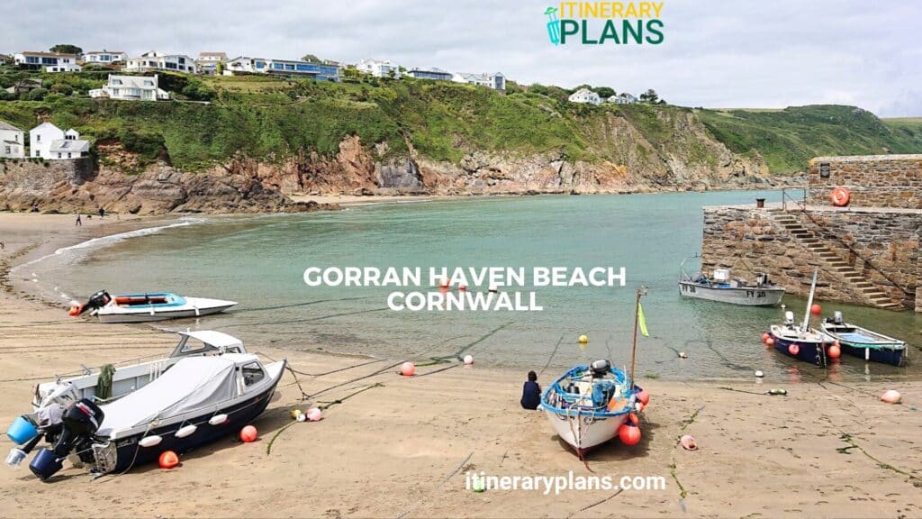 View of Gorran Haven Beach Cornwall, featuring moored boats on sandy shores, calm turquoise waters, and cliffs in the background.