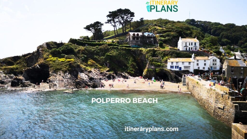 Scenic view of Polperro Beach in Cornwall, featuring a small sandy cove surrounded by rocky cliffs, traditional whitewashed houses, and lush greenery under a clear blue sky.