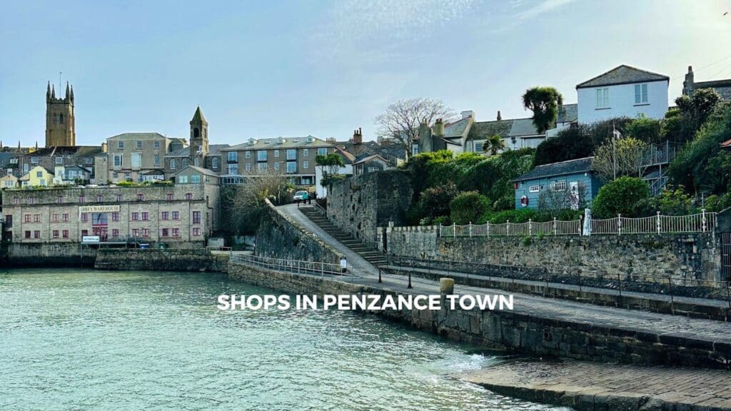 View of charming shops and historic buildings in Penzance Town, with a coastal walkway and lush greenery in the background.