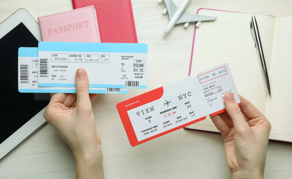 Two hands holding flight tickets over a table with passports, a tablet, a notebook, a pen, and a toy airplane, evoking a sense of travel excitement.
