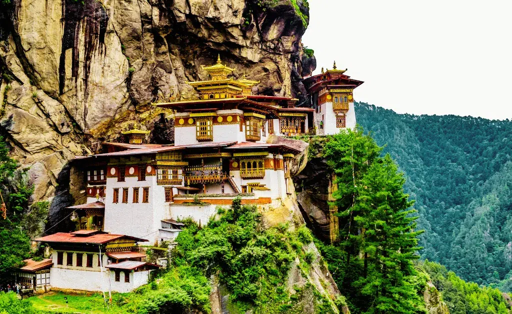 A majestic monastery perched on a rugged cliffside, surrounded by lush green forest. The structure features intricate golden roofs, evoking a serene, spiritual atmosphere.