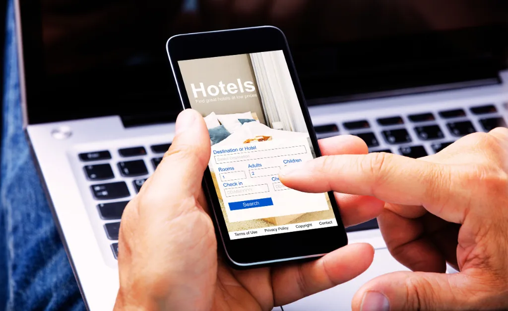 A person uses a smartphone app to book a hotel, with a laptop in the background. The screen shows a search form for destinations, dates, and guest details.