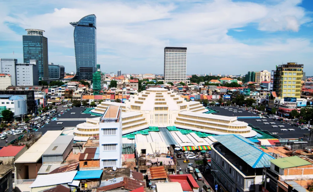 Aerial view of a city with a prominent central market building featuring a beige, art deco design. Surrounding it are modern skyscrapers and a bustling urban landscape under a blue sky with scattered clouds.