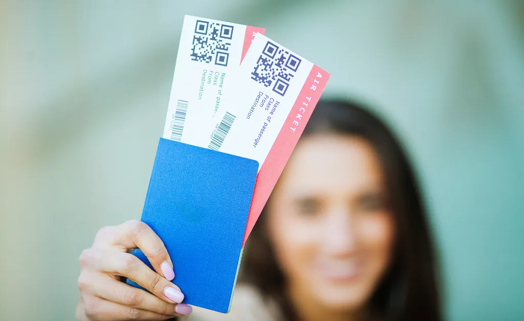 A woman holds up two airline tickets with QR codes and a blue passport blurred in the background. The image conveys excitement and anticipation for travel.
