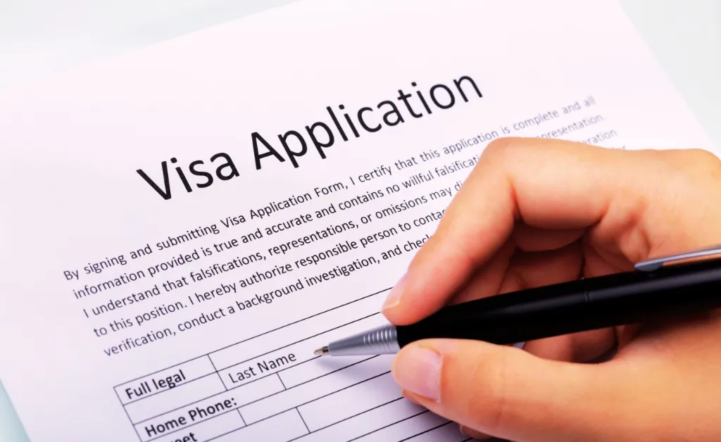 A hand holding a pen is filling out a printed visa application form.