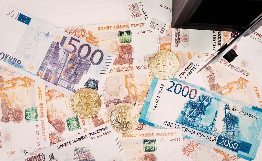 Euro and Russian ruble banknotes, bitcoin coins, and a pen beside an open wallet create a mixed currency and finance theme.