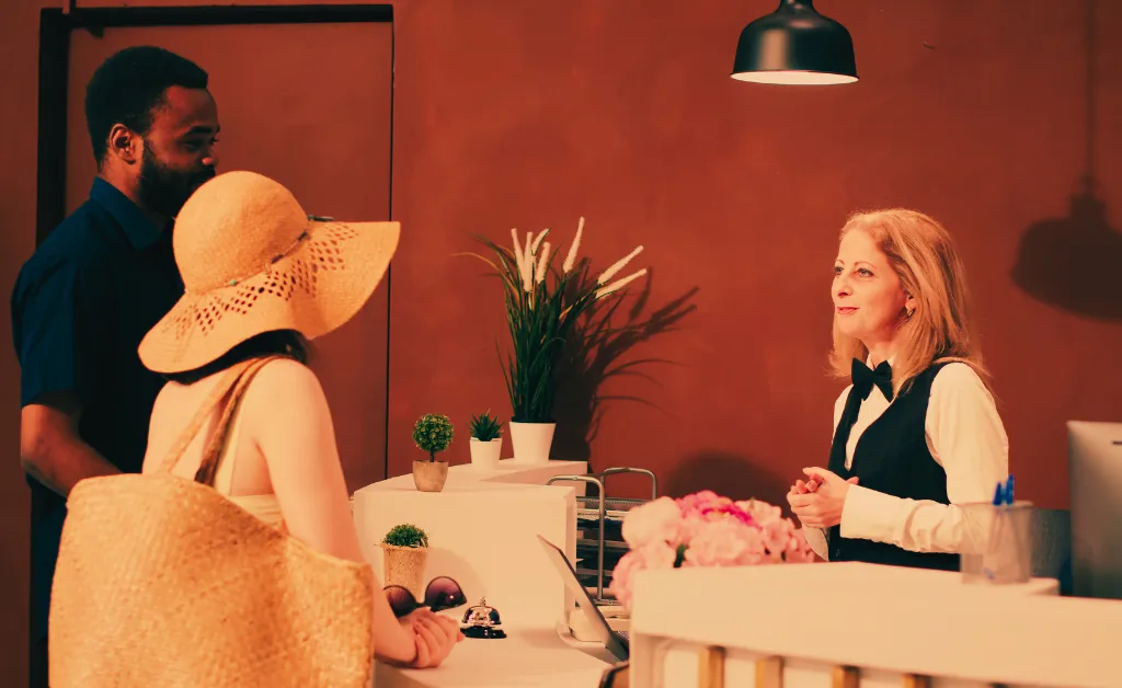 A couple at a hotel reception desk, warmly greeted by a smiling receptionist. The woman wears a sun hat, evoking a casual, welcoming atmosphere.