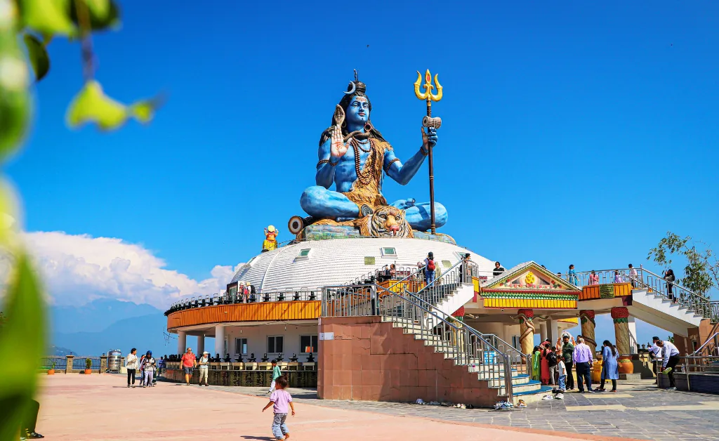 A large blue statue of a deity holding a trident sits atop a tiered structure under a clear blue sky. Tourists explore the area, creating a lively atmosphere.