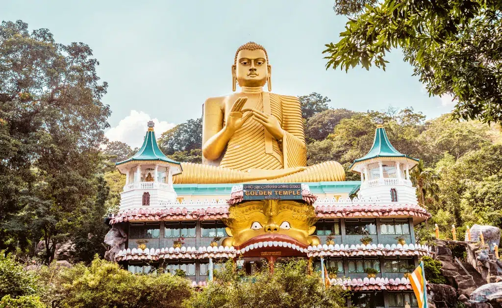 A large golden Buddha statue sits atop a richly decorated temple with ornate details and vibrant colors, surrounded by lush green trees and clear skies.