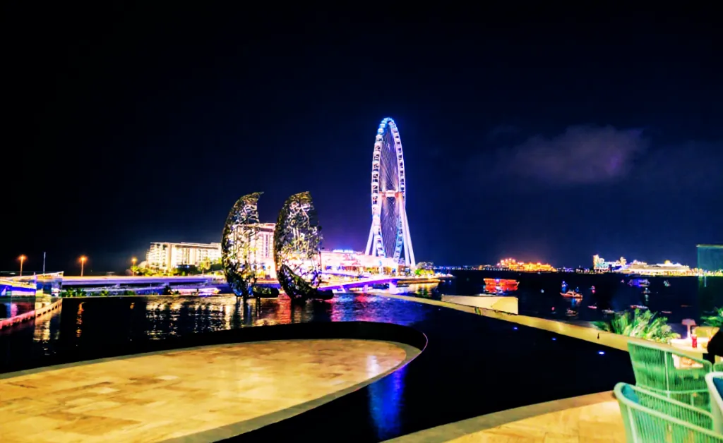 Nighttime cityscape with a glowing Ferris wheel and artistic twin sculptures in the foreground. Bright lights reflect on the water, creating a vibrant, lively atmosphere.