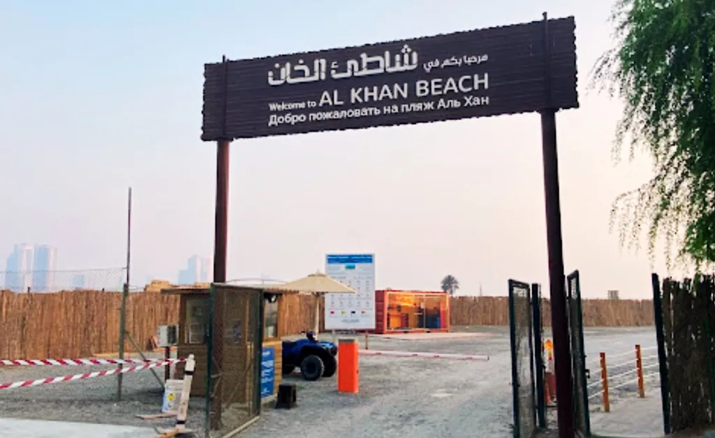 Entrance to Al Khan Beach, marked by a wooden sign in English, Arabic, and Russian. Surrounding area features sandy terrain and fencing.