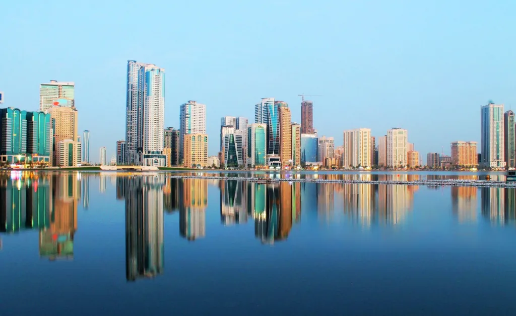 A city skyline with modern high-rise buildings is reflected in calm water under a clear blue sky, conveying serenity and urban elegance.