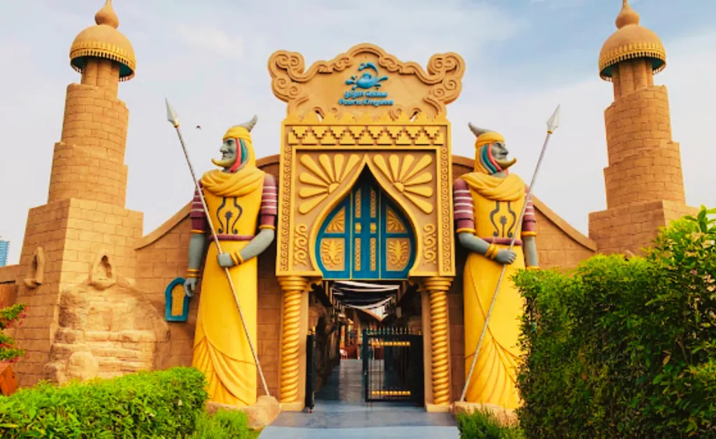 Ornate entrance with two tall, colorful guardian statues in yellow robes holding spears. Sandstone towers flank the doorway, evoking an ancient architectural style.