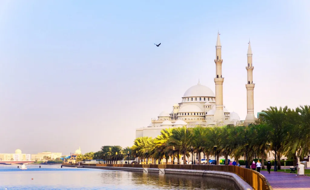 A majestic mosque with tall minarets stands beside a serene waterfront lined with palm trees, under a clear sky, evoking a sense of tranquility.