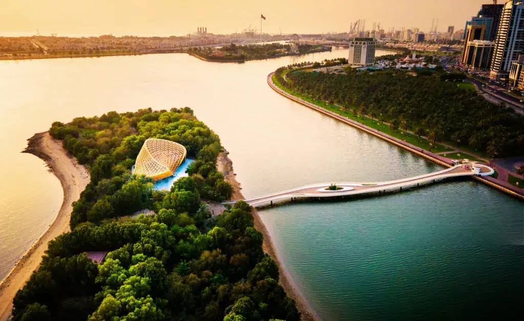 Aerial view of a lush, green island with a geometric pavilion near a serene river. A curved bridge connects the island to the mainland under a golden sunset.