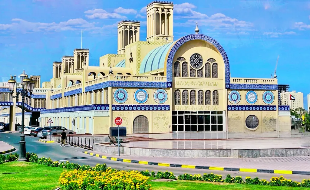 A vibrant Middle Eastern market building with blue tile details and arched windows under a clear sky. Lush greenery and a road in the foreground.