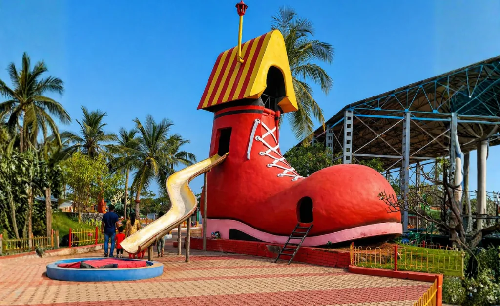A large, whimsical red boot-shaped playground structure with a yellow slide and a small ladder. Surrounding it are palm trees and a group of children playing, conveying a joyful, playful atmosphere.