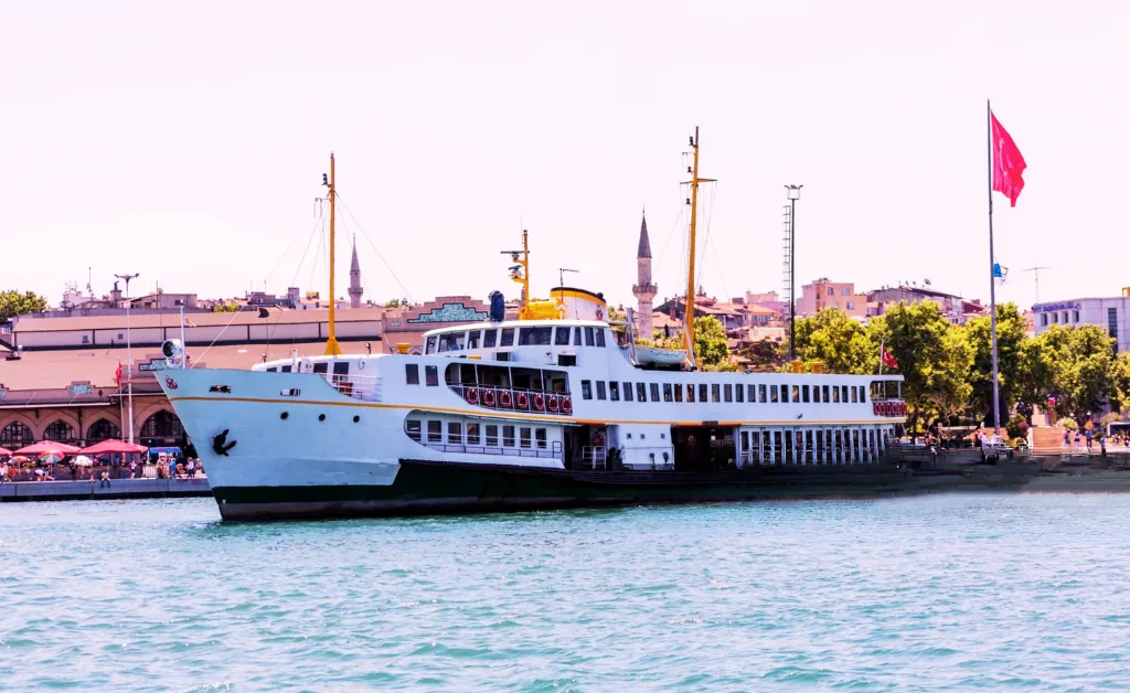A white ferry with yellow masts docked at a city port with a large red flag. The background features city buildings and a minaret under a clear sky.