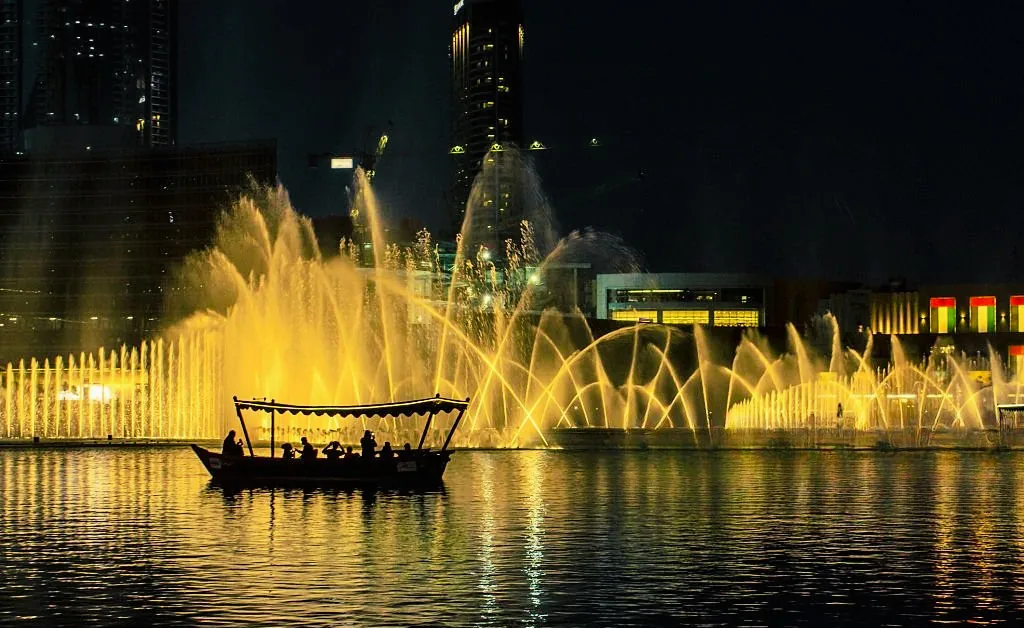 Silhouette of a boat with passengers on a lake at night, set against a backdrop of illuminated fountains spraying water in golden arcs.