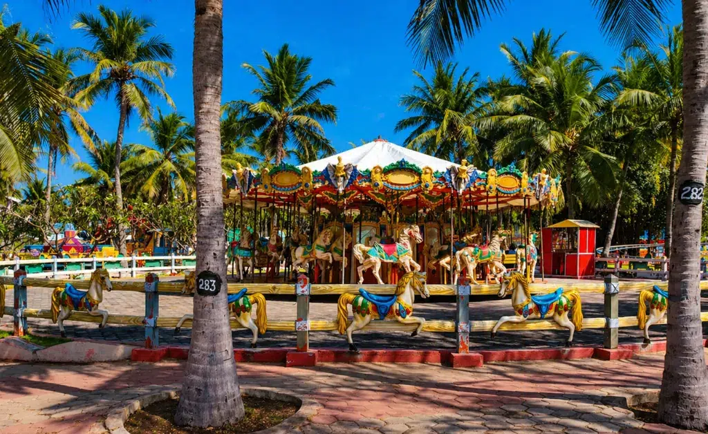 A colorful carousel sits amidst tropical palm trees under a clear blue sky. Brightly painted horses add a joyful, nostalgic feel to the vibrant scene.