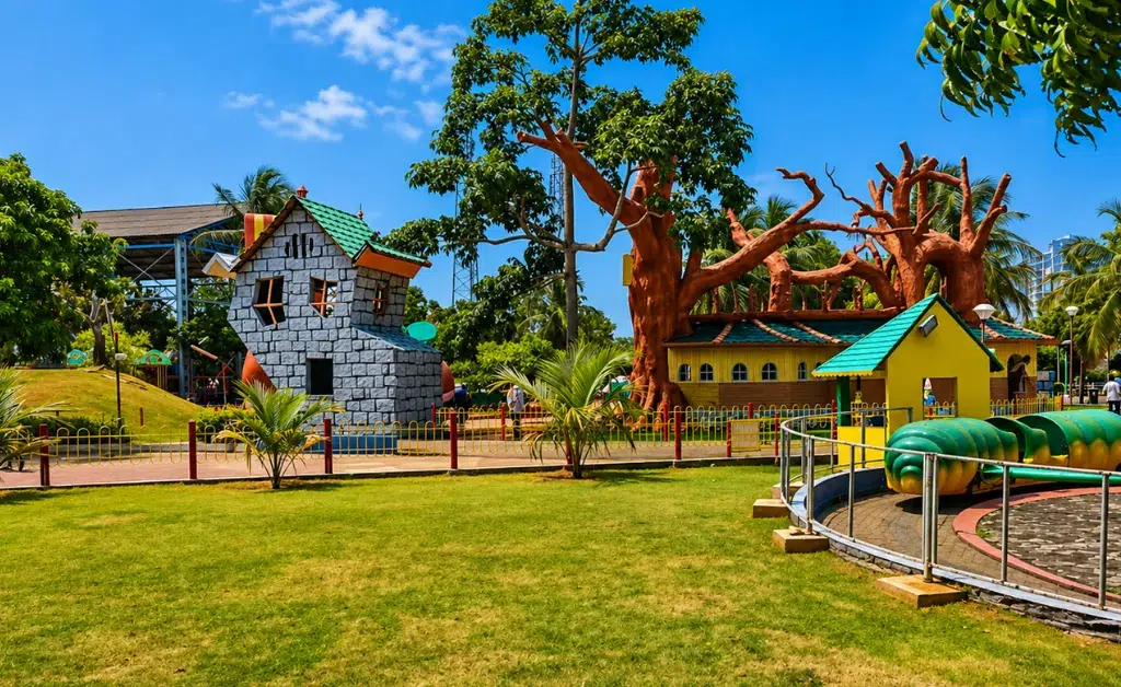 A vibrant amusement park scene with whimsical buildings, including a stone house and tree-like structures, set against a clear blue sky and lush greenery.