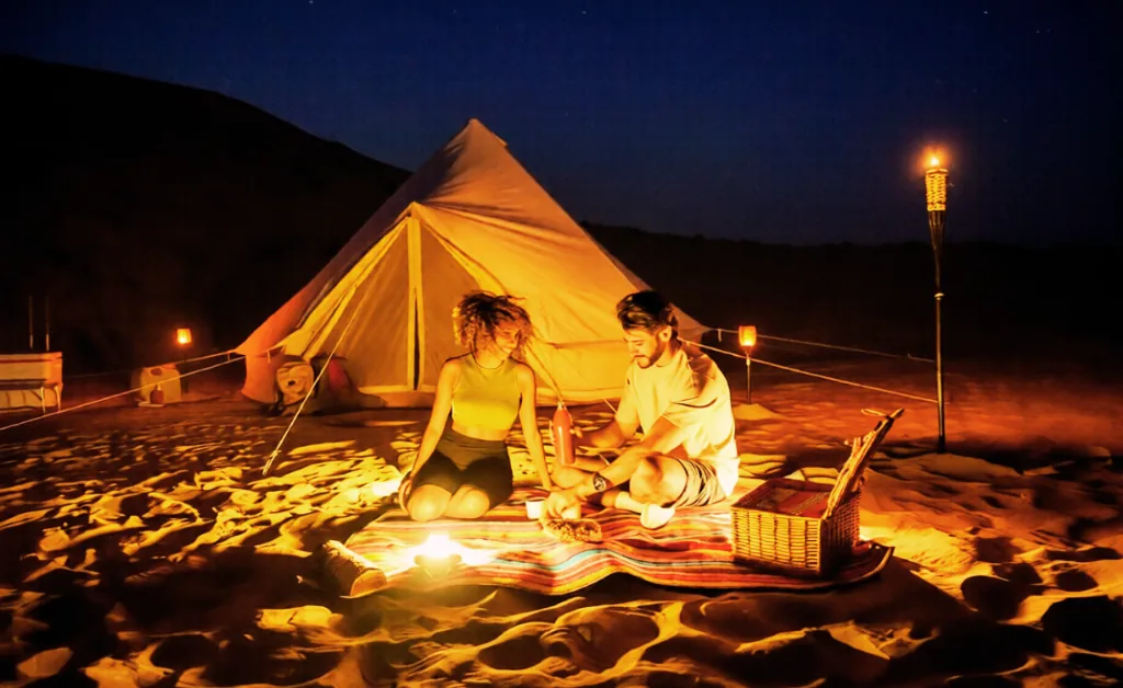 A couple sits on a striped blanket in front of a lit tent in the desert at night. Surrounded by lanterns and torches, the scene feels warm and intimate.