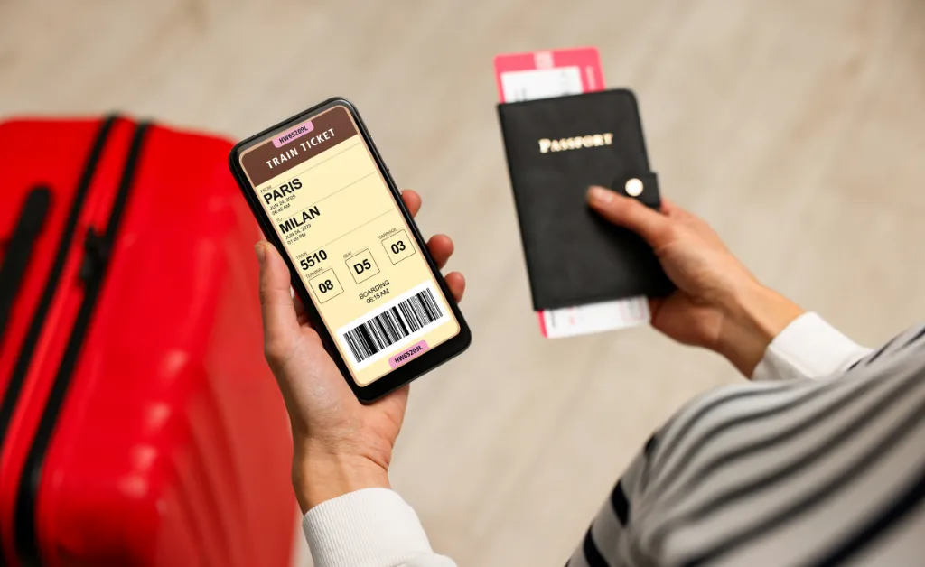 A person holds a smartphone displaying a train ticket from Paris to Milan, a passport, and a boarding pass, next to a red suitcase, suggesting travel plans.