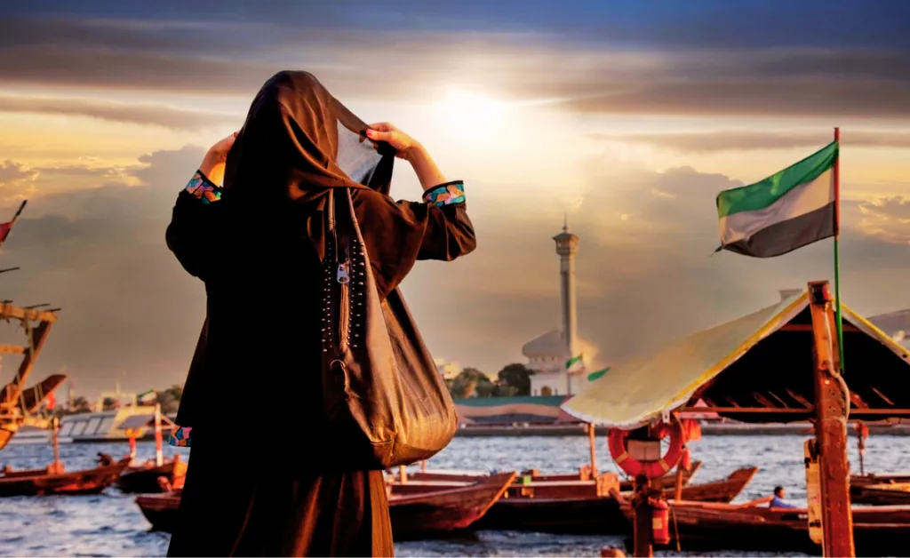 A woman in a black abaya stands by the water at sunset, facing a distant mosque. Boats and an Emirati flag are visible, conveying a serene ambiance.