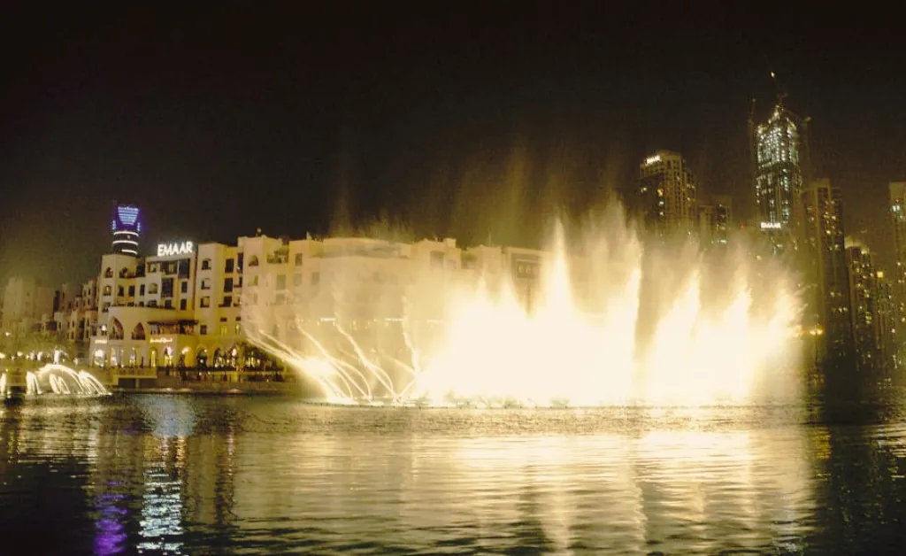 Illuminated fountains at night, spraying water high into the air with cityscape and buildings in the background, creating a magical, vibrant scene.