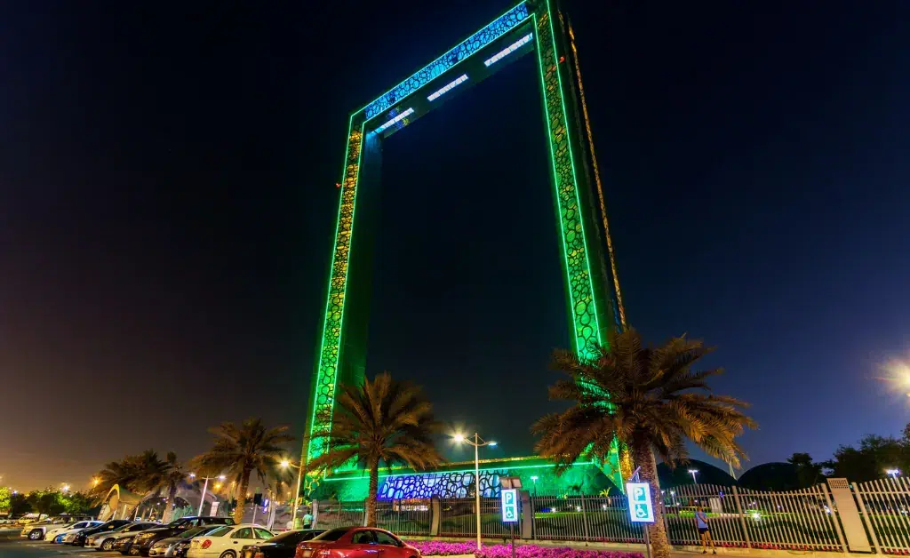 The image shows the Dubai Frame at night, illuminated in vibrant green lights. Palm trees are in the foreground, creating a striking architectural scene.