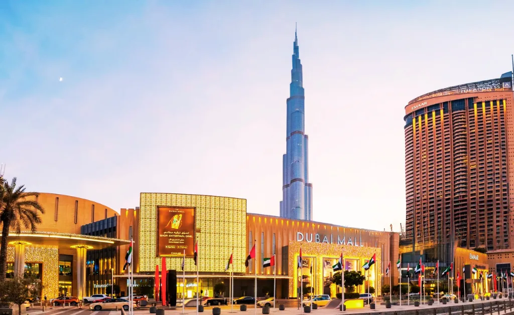 Dubai Mall facade illuminated at sunset with the Burj Khalifa towering in the background. Flags line the foreground, creating a vibrant, bustling atmosphere.