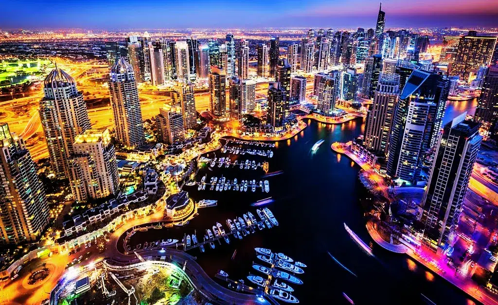 Vibrant cityscape of Dubai Marina at night, showcasing illuminated skyscrapers, a dark water canal with boats, and bustling streets, exuding a lively urban atmosphere