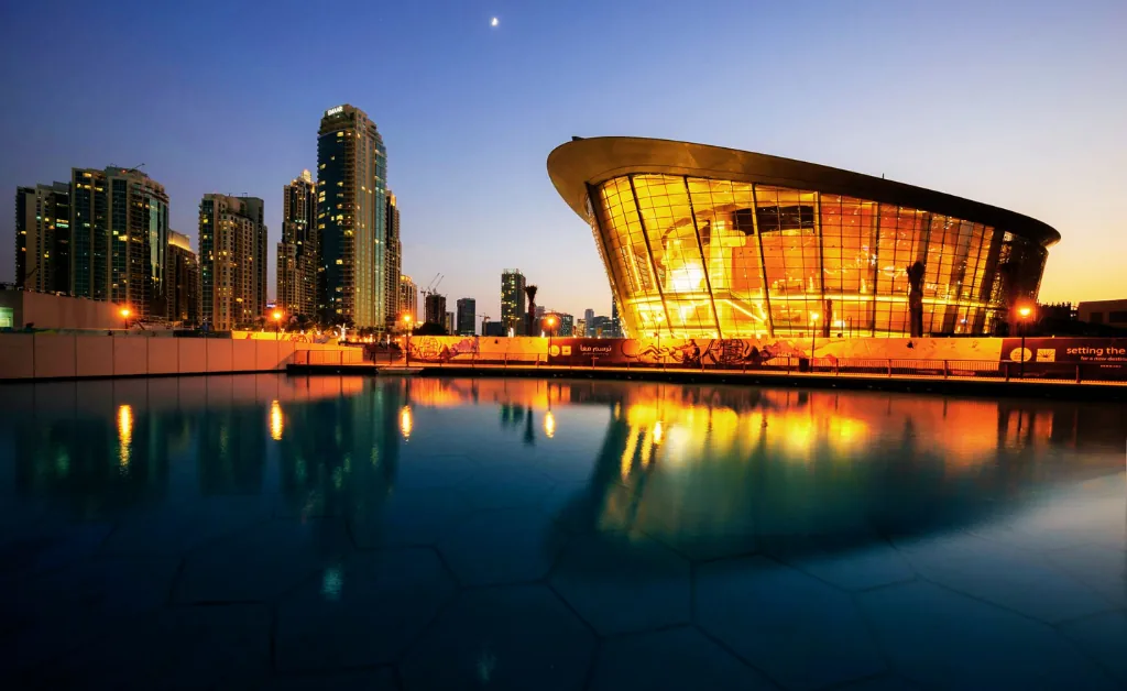 Cityscape at dusk with a futuristic, glowing building reflecting on water. High-rise skyscrapers line the background under a deep blue sky with a visible moon.