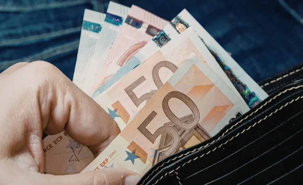 A hand holding several euro banknotes, including 50 denominations, near a striped wallet. Denim fabric in the background. The scene conveys monetary value.