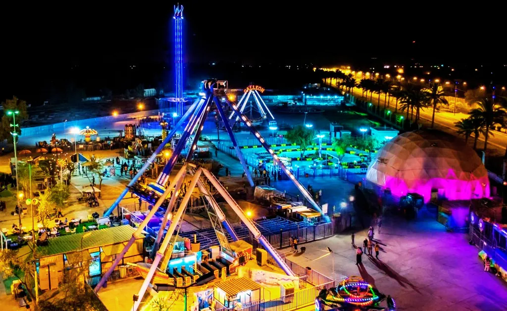 A vibrant nighttime amusement park, illuminated with colorful lights. Rides and attractions, including a Ferris wheel, create a lively, festive atmosphere.