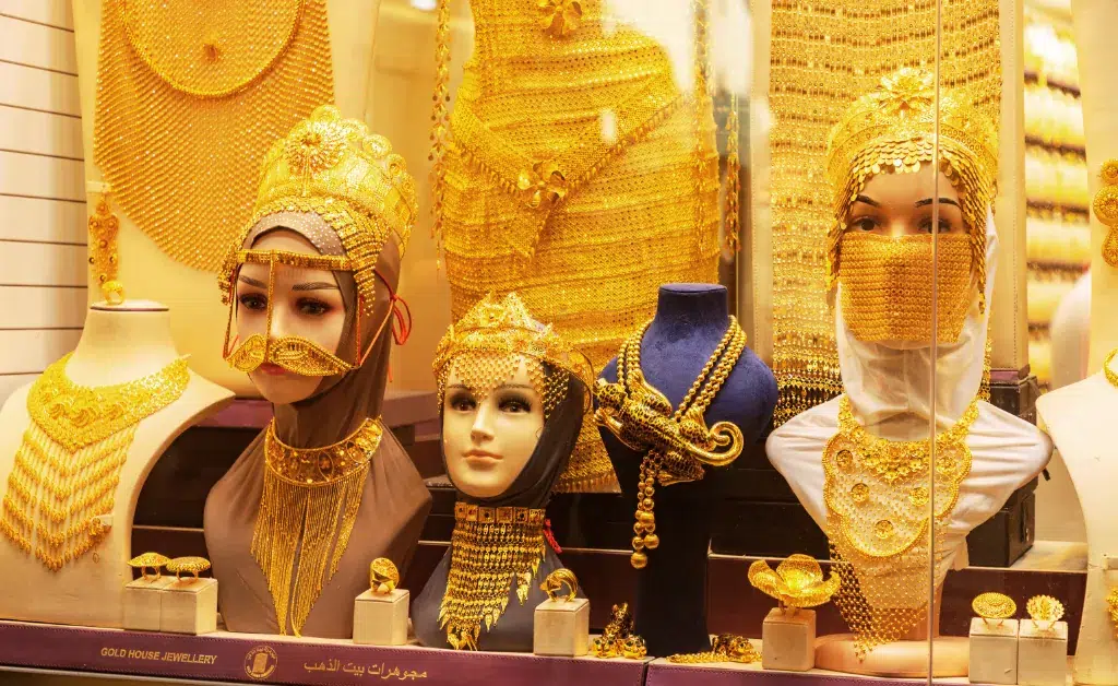 Window display showcasing intricate gold jewelry sets on mannequins, featuring ornate headpieces and necklaces. The scene exudes luxury and opulence.