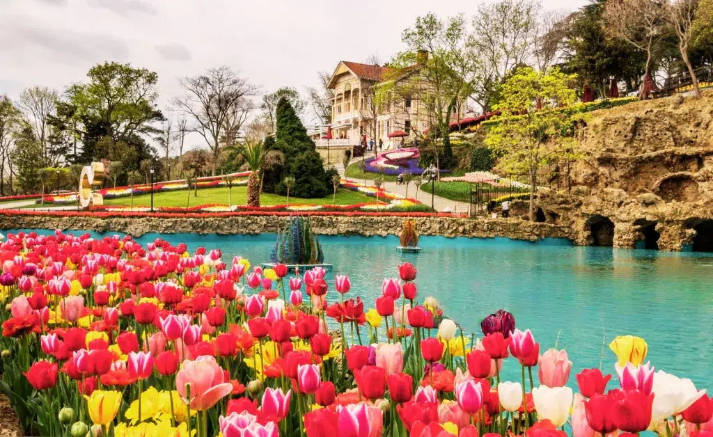 A vibrant garden with colorful tulips borders a turquoise lake. A historic building stands on a hill, surrounded by lush greenery and pathways.
