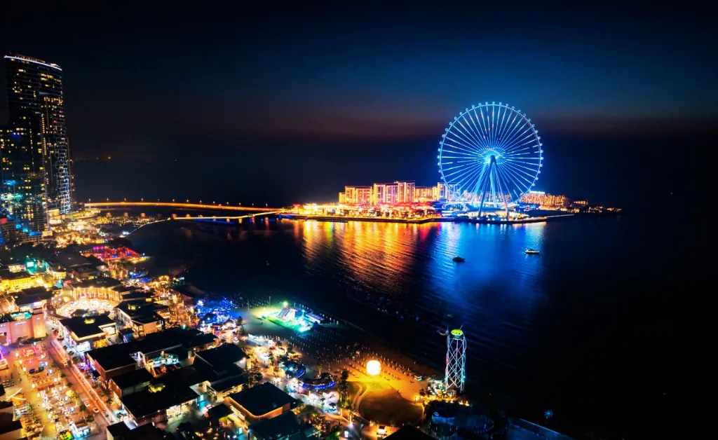 A vibrant night scene of a city with a brightly lit Ferris wheel reflecting on water. Colorful lights illuminate the bustling area, conveying a lively atmosphere.