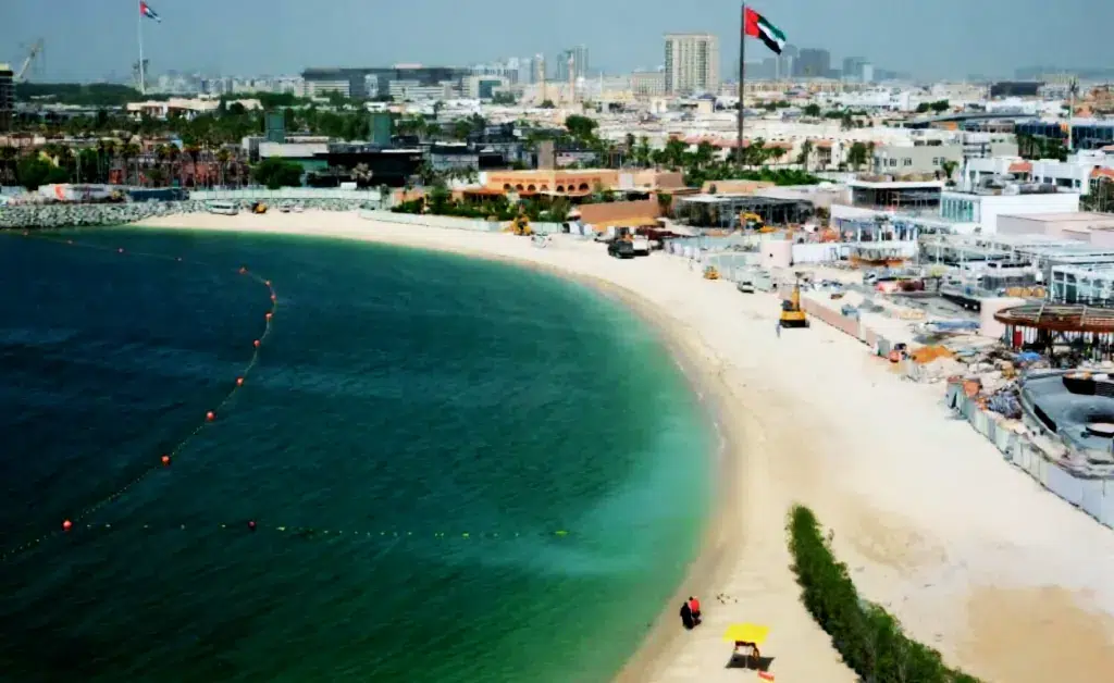 Aerial view of a sunny beach with turquoise water and a sandy shore. A cityscape with a prominent flagpole is visible in the background. Peaceful ambiance.