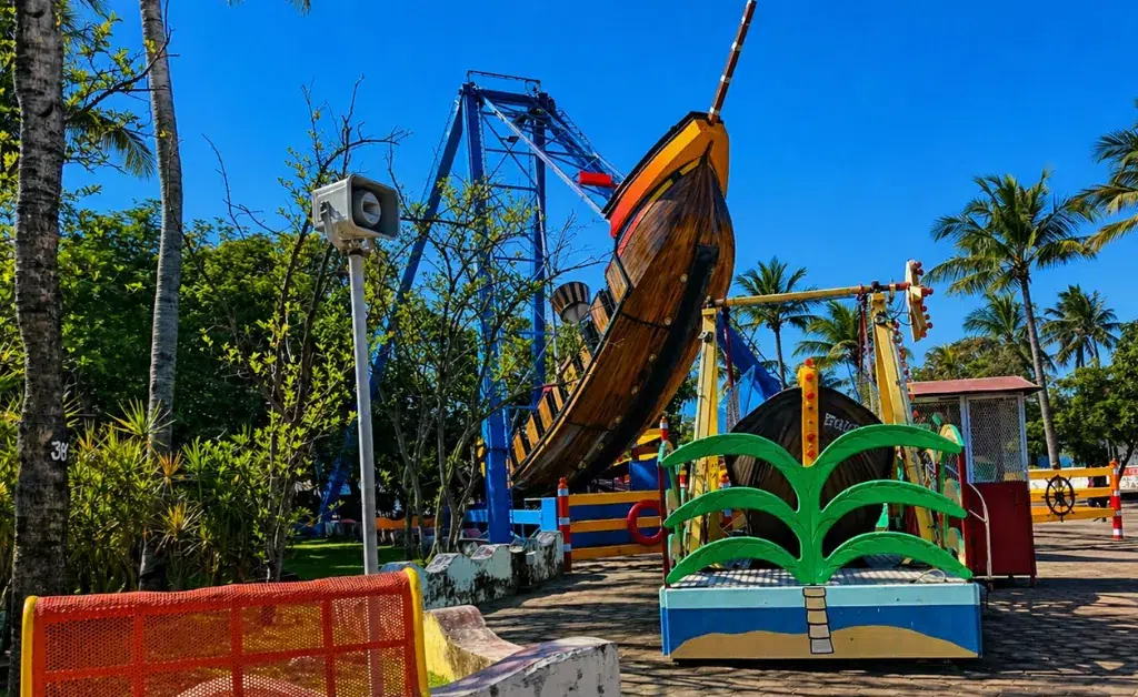 A carnival ride shaped like a pirate ship is swinging under a clear blue sky. Palm trees and colorful benches surround the lively amusement park scene.