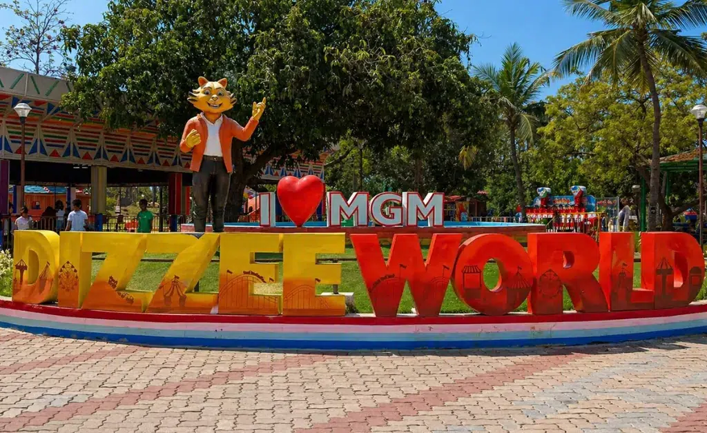 Entrance to a theme park featuring a colorful "Dizzee World" sign with a cheerful cat mascot. Lush greenery and palm trees create a fun, inviting atmosphere.