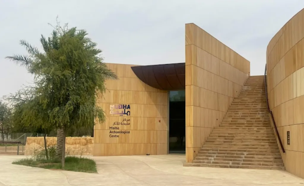 Modern building facade of the Mleiha Archaeological Centre with sleek wooden panels, an adjacent tree, and a wide staircase leading to an upper level.