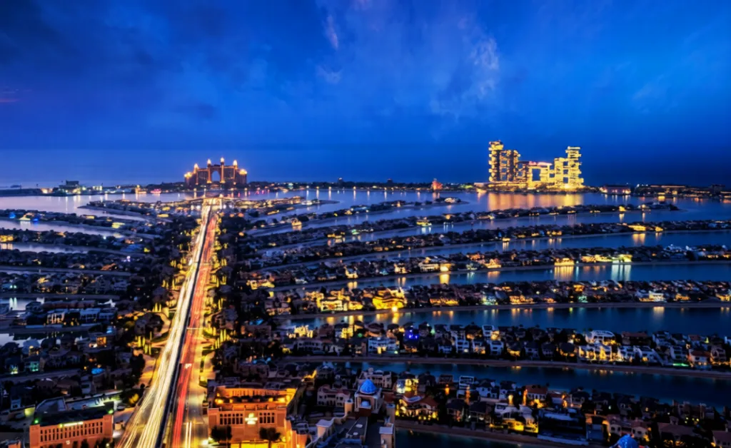 Aerial view of a palm-shaped island at night, illuminated by vibrant city lights. Skyscrapers are visible in the distance against a dark blue sky.
