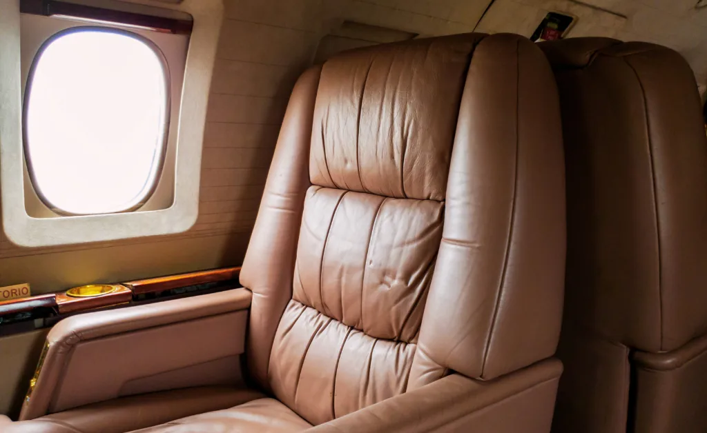 Luxurious brown leather airplane seat beside a window, with a wooden trim and cup holder on the side. The ambiance conveys comfort and opulence.