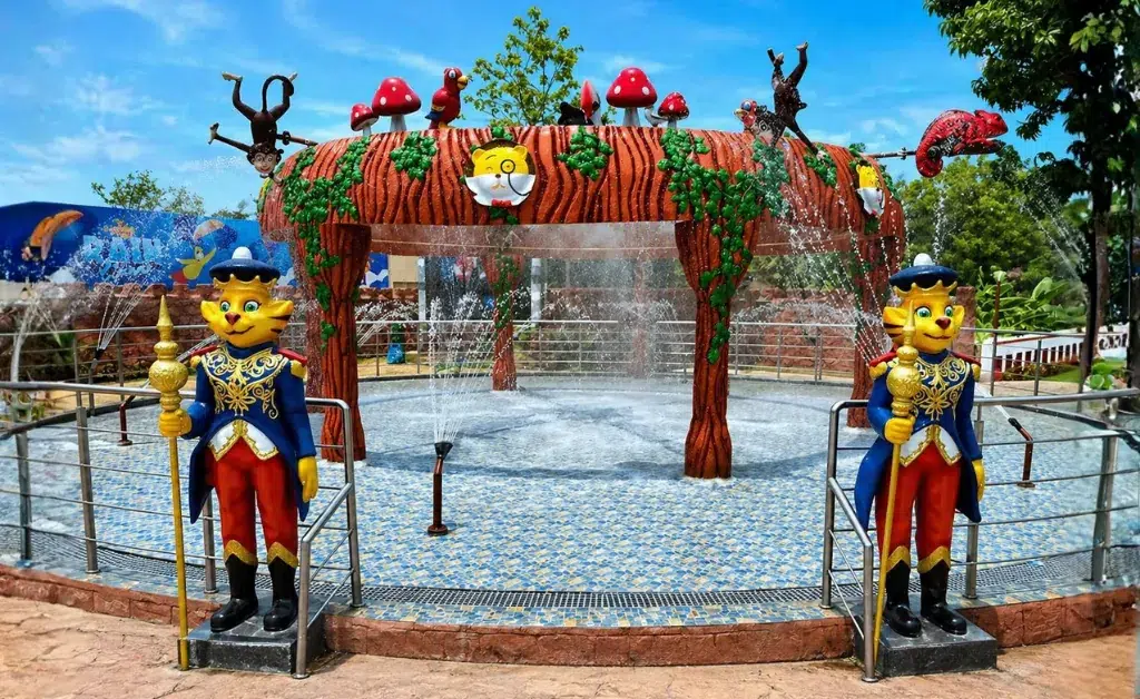 Fun water play area with mushroom decorations and tree-like arches spraying water. Two cat statues in uniform stand at the entrance, under a clear sky.
